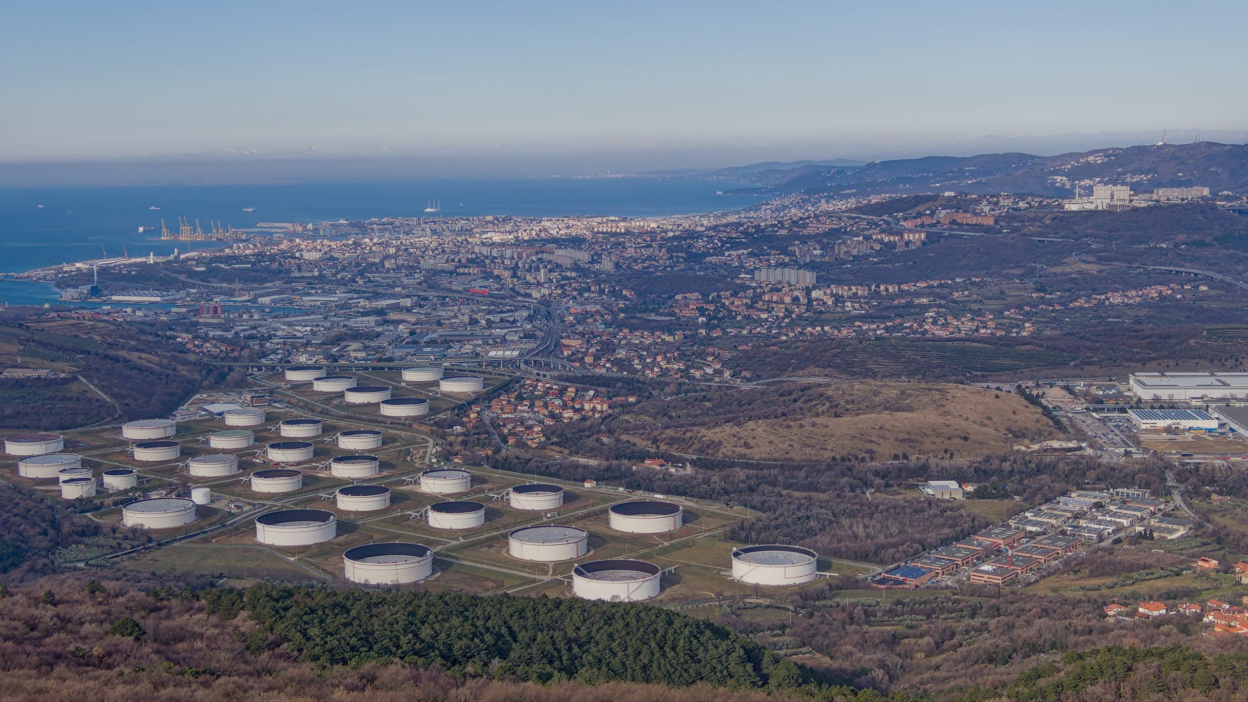 Aerial view of a coastal petroleum tank farm, with a commercial port and open sea beyond it.