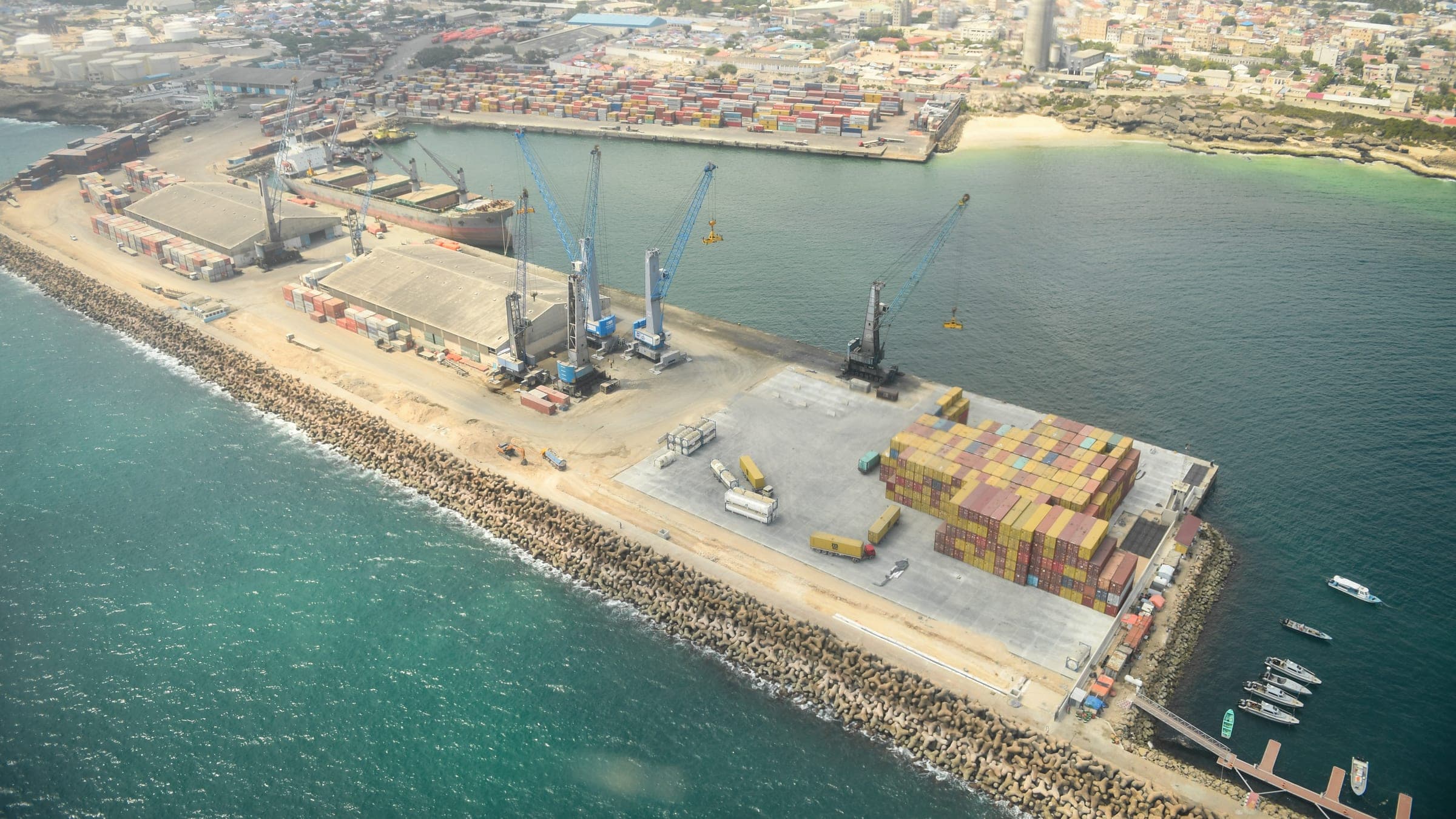 Aerial view of a working seaport, showing quayside cranes, stacked containers and a breakwater.